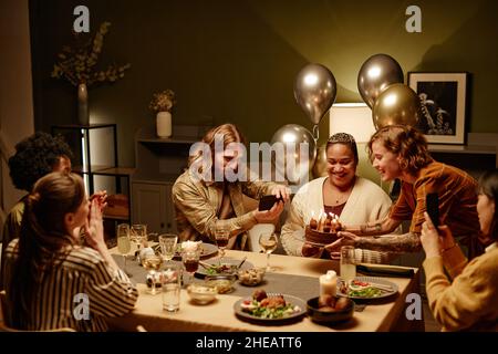 Eine Gruppe glücklicher junger Freunde, die gemeinsam am Esstisch im Zimmer Geburtstag feiern, Fotos machen und Geburtstagskuchen blasen Stockfoto