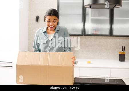 Multirassische Frau Auspacken Paket Karton mit Online-Bestellung. Zufriedene Biracial junge Frau Auspacken Paket-Box, während sie in der Küche zu Hause stehen. Aufgeregte Frau, die die gelieferten Waren ansieht Stockfoto