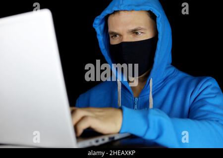 Mann in Maske und blauem Hoodie mit Laptop auf schwarzem Hintergrund. Konzept von Cyberkriminalität, Hacking und Technologie Stockfoto