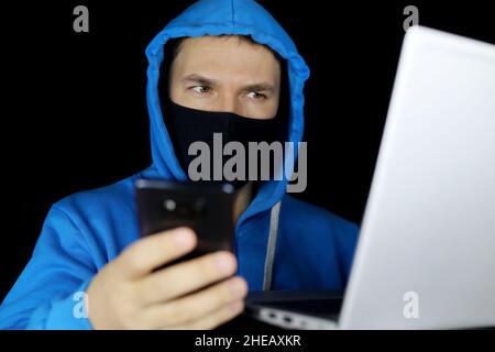 Mann in Maske und blauem Hoodie sitzend mit Smartphone in der Hand am Laptop auf schwarzem Hintergrund. Konzept von Cyberkriminalität, Hacking und Technologie Stockfoto