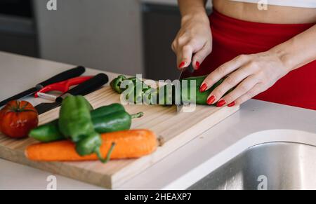 Unverkennbare Hausfrau mit roten Nägeln hackt reifen grünen Pfeffer auf Schneidebrett in der Nähe von Tomaten und Karotten, während sie gesundes Gericht zu Hause zubereitet Stockfoto