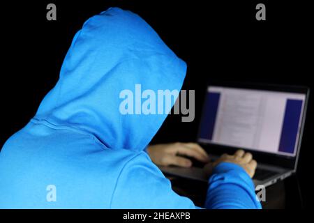 Cyberkriminalität, Hacking und Technologiekriminalität. Mann im blauen Hoodie sitzt mit Laptop auf schwarzem Hintergrund, Konzept der Torrent-Piratentum und illegale Inhalte Stockfoto