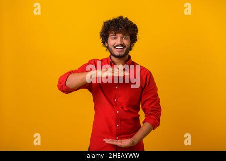 Hübscher indischer Mann mit Bart, der auf orangefarbenem Hintergrund steht und mit Händen gestikelt, während er ein großes und großes Zeichen zeigt, ein Maßsymbol. Messkonzept Stockfoto