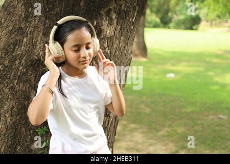 Fröhliches junges indisches Mädchen, das mit dem Kopfhörer Musik im Grün hört. Sie ist umgeben von grüner Umgebung, verloren in der Musik Stockfoto