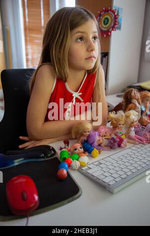 Ein sechsjähriges Mädchen, das am Computer zu Hause liest, begleitet von zahlreichen Puppen Stockfoto