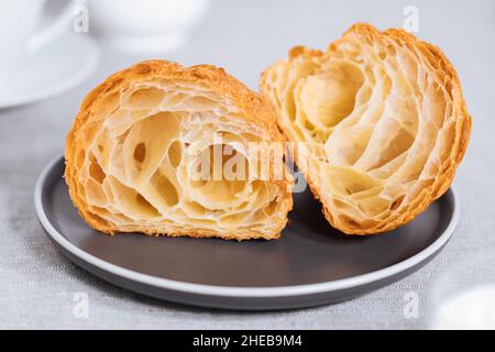 Croissant in hellgrauer Umgebung und in zwei Stücke geschnitten, mit einer Tasse Kaffee und Milch und anderen Croissants in verschwommenem Hintergrund. Einfach und elegant b Stockfoto