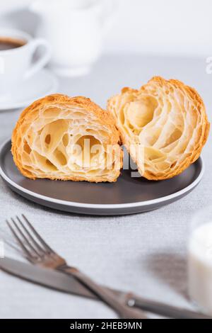 Croissant in hellgrauer Umgebung und in zwei Stücke geschnitten, mit einer Tasse Kaffee und Milch und anderen Croissants in verschwommenem Hintergrund. Einfach und elegant b Stockfoto