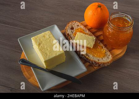 Gesundes Frühstück mit rustikalem Brot, süßer Orangenmarmelade und Bio-Butter auf einem rustikalen Holztablett auf einem Tisch mit einem Spread-Messer Stockfoto