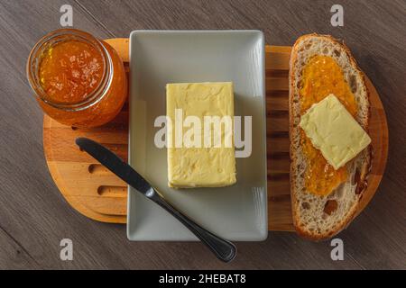 Gesundes Frühstück mit rustikalem Brot, süßer Orangenmarmelade und Bio-Butter auf einem rustikalen Holztablett auf einem Tisch mit einem Spread-Messer Stockfoto