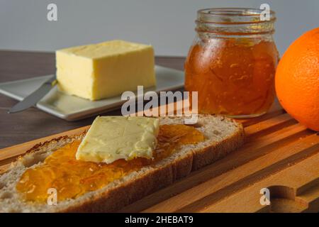 Rustikales Brot mit Orangenmarmelade und Butter auf einem Holzbrett mit einem Glas Marmelade im Hintergrund neben der Butter Stockfoto