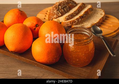 Natürliche süße Orangen, um Marmelade in einem Behälter mit rustikalem Brot im Hintergrund und einem Esslöffel, um ein reichhaltiges Frühstück vorzubereiten Stockfoto