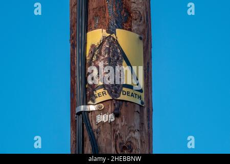 Stark verbranntes dreieckiges Warnschild „Danger of Death“, das auf einem Holzmast mit vertikalen Kabeln vor blauem Himmel angebracht ist Stockfoto