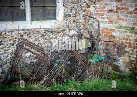 Ein kleiner Stapel rostender, metallgerahmter Stühle mit fehlender oder zerrissener Leinwand an einer Feuerstein-, Backsteinmauer und einem Pfosten-Fenster Stockfoto