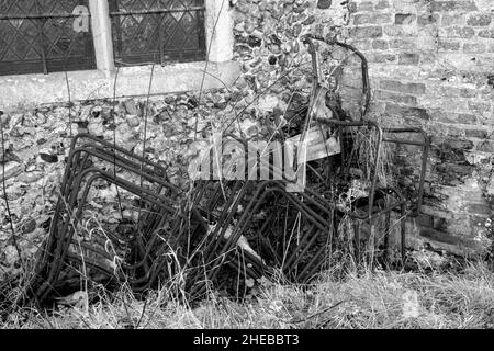 Ein kleiner Stapel rostender, metallgerahmter Stühle mit fehlender oder zerrissener Leinwand an einer Feuerstein-, Backsteinmauer und einem Pfosten-Fenster Stockfoto