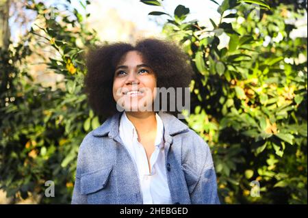Mode close up stilvolles Porträt der attraktiven jungen natürlichen Schönheit afroamerikanische Frau mit afro Haar in blauem Mantel und weißem Hemd posiert im Freien. Glückliche Dame lacht mit perfektem Lächeln und Zähnen Stockfoto