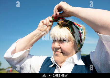 Eine ältere weißrussische oder ukrainische Frau bindet ein ethnisches Kopftuch auf ihren Kopf. Stockfoto