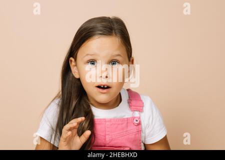 Porträt eines Kindes mit überraschten runden Augen, leicht geöffnetem Mund und aufgehender Hand, die die Kamera in einem leuchtend rosa Jumpsuit und einem weißen T-Shirt anschaut Stockfoto