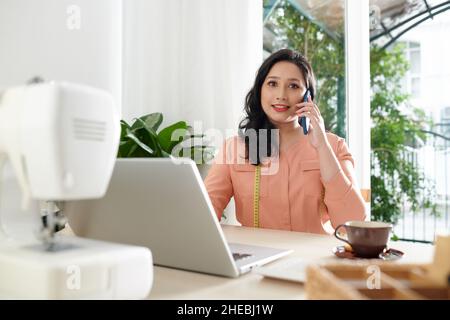 Porträt einer lächelnden Näherin, die auf dem Smartphone mit der Kundin spricht und in ihrem Arbeitsatelist am Laptop arbeitet Stockfoto