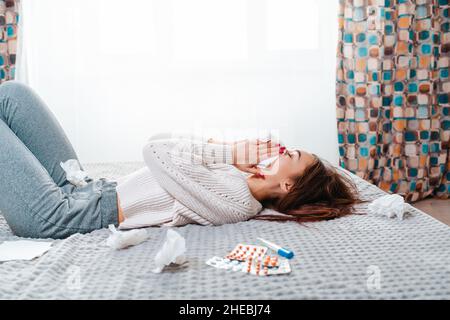 Das Konzept von Krankheiten und saisonalen Erkältungen. Eine Brünette liegt auf dem Bett und bläst ihre Nase in ein Taschentuch. Neben ihr sind Pillen, ein thermometer Stockfoto