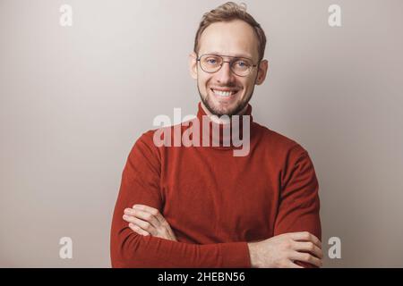Porträt eines Mannes mit Brille auf grauem Hintergrund. Glücklicher Kaukasischer in rotem Rollkragen, der die Kamera über einer grauen Wand mit Platz für Kopien ansah Stockfoto
