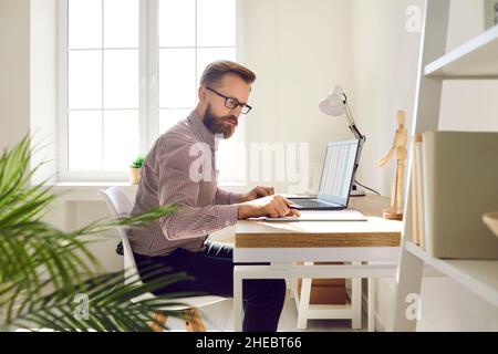 Fokussierter Buchhalter, der mit Papierdokumenten und Software für elektronische Rechnungen auf einem Laptop arbeitet. Stockfoto
