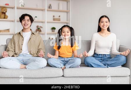 Lächelnde asiatische Familie sitzt in Lotusposition, üben Yoga auf dem Sofa in minimalistischer Wohnzimmereinrichtung zur freien Zeit Stockfoto