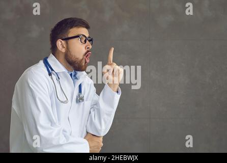Der junge Arzt, der Chirurg, zeigt den Finger nach oben und sieht inspiriert von genialen Gedanken aus Stockfoto