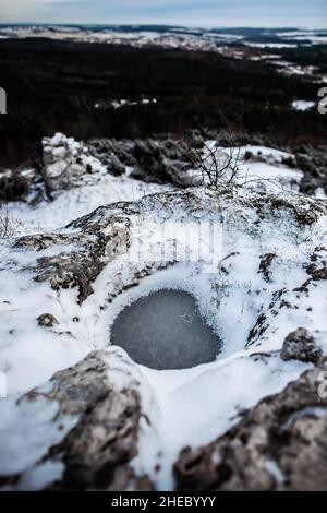 Kleine gefrorene Pfütze auf dem Gipfel des flachen Gipfels auf dem Hügel in der Nähe des Winters, wunderschöne schneebedeckte Winterlandschaft mit Felsen und Wald im Hintergrund Stockfoto