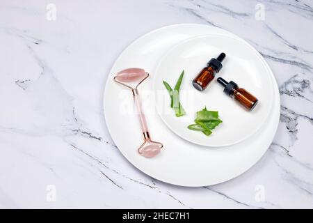 Weiße Teller mit Aloe Vera-Blättern, Hautpflegeöl oder Serum und Gesichtsmassagewoller auf weißem Marmorboden. Natürliches Hautpflege- und Spa-Konzept. Stockfoto
