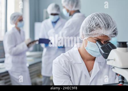 Junge Wissenschaftlerin in Schutzmaske sitzt am Tisch und schaut durch das Mikroskop, sie arbeitet im Labor Stockfoto