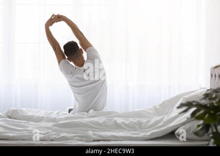 Mann, Der Im Bett Sitzt Und Die Arme Nach Dem Aufwachen Dehnt, Rückansicht Stockfoto