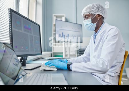 Afrikanischer Chemiker in Maske sitzt an seinem Arbeitsplatz vor dem Computermonitor und tippt auf der Tastatur und arbeitet im Büro mit Charts Stockfoto