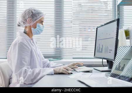 Junge Ärztin mit Maske und Handschuhen, die am Tisch vor dem Computermonitor sitzt und im Büro mit Charts arbeitet Stockfoto