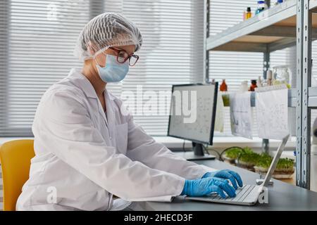 Junge Forscherin in Maske und Handschuhen konzentriert sich auf ihre Online-Arbeit am Laptop im Labor Stockfoto