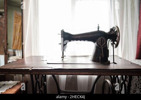 Vintage mechanische Nähmaschine mit Tisch steht an einem Fenster in einem rustikalen Haus. Stockfoto