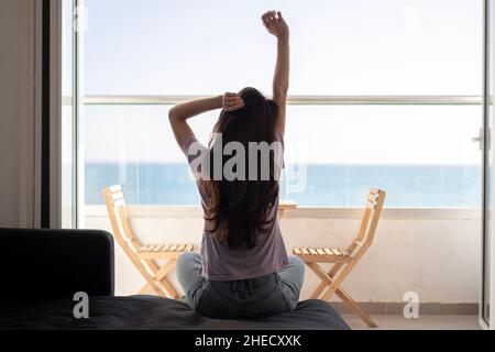 Eine junge Frau sitzt auf dem Sofa in der Nähe des Balkons und dehnt sich am Morgen Stockfoto