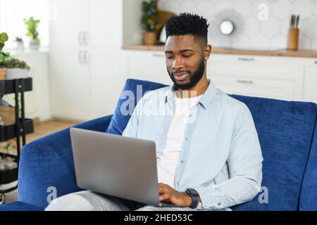 Lächelnder afroamerikanischer Freiberufler benutzt Laptop-Computer, der zu Hause auf der Couch sitzt, hindu-Typ genießt Fernarbeit von zu Hause aus in gemütlicher Atmosphäre. Schwarzer Mann verbringt Freizeit online Stockfoto