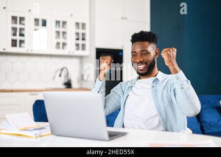 Großartige Jobchancen. Aufgeregt guckt auf Laptop-Bildschirm und schreiend vor Freude, erhielt Post, gute Nachrichten, Afrikaner feiern Sieg oder bekam einen Job, Heben Fäuste sitzen am Schreibtisch zu Hause Stockfoto