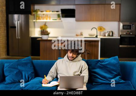 High-Angle-Ansicht des multiethnischen Hindu-Freiberuflers, der einen Laptop für die Arbeit oder das Studium in der Ferne von zu Hause aus verwendet. Konzentrierter indischer Mann, der sich das Online-Webinar ansieht und sich Notizen im Notizbuch ansieht Stockfoto