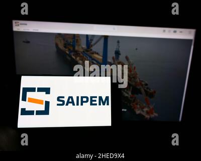 Person, die Mobiltelefon mit dem Logo des italienischen Oilfield-Dienstleisters Saipem S.p.A. auf dem Bildschirm vor der Webseite hält. Konzentrieren Sie sich auf die Telefonanzeige. Stockfoto