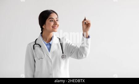 Fröhliche tausendjährige nette indische Dame Arzt in Uniform schreibt auf virtuellen Brett in Luft Stockfoto