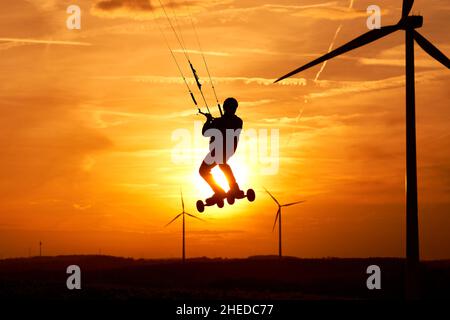 Kite Land Boarder springt vor der untergehenden Sonne. 1 Mann bei einer extremen Sportart in Aktion mit einem Kiteboard. Landschaft mit Windturbinen als Silhouette. Stockfoto