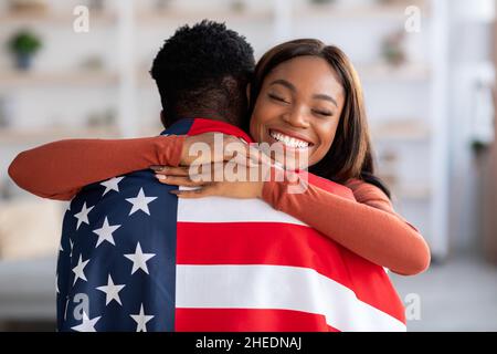 Unabhängigkeitstag. Glückliche Schwarze Frau Umarmt Ehemann In American Flag Eingewickelt Stockfoto