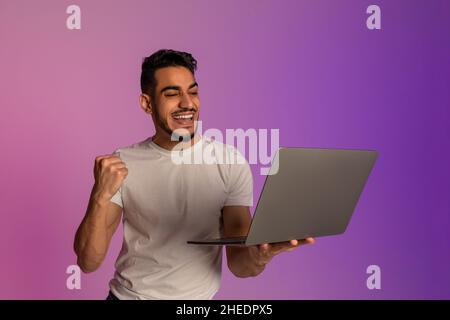 Cooler junger arabischer Mann mit Laptop, der YES-Geste macht, begeistert über riesigen Online-Lotteriegewinn oder Erfolg im Neonlicht Stockfoto