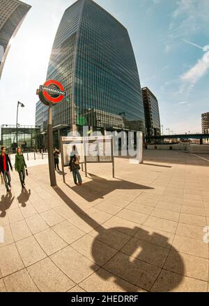 London, Großbritannien - 06. April 2007: Sonne scheint auf unterirdisches Schild am Bahnhof Canary Wharf, vorbeigehende Menschen, hohe Wolkenkratzer im Hintergrund - w Stockfoto