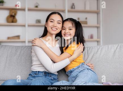 Zufriedene, hübsche, tausendjährige chinesin und Teenager-Mädchen umarmen sich und sitzen auf dem Sofa im Wohnzimmer Stockfoto