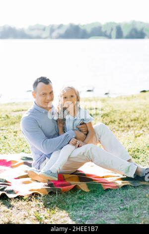 Ein glücklicher Vater hält seine Tochter in den Armen. Sie haben Spaß in der Natur, sitzen im Sommer auf einer Decke am See Stockfoto