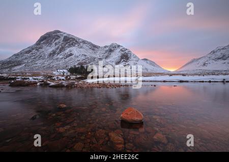 Eine abgelegene Hütte und ein ruhiger Fluss mit Stein im Vordergrund am Fuße des Buachaille Etive Mor am Eingang zum Tal von Glencoe in den schottischen Highlands, Schottland Stockfoto