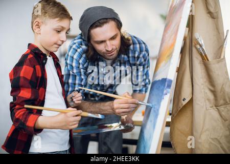 Fokussierter kaukasischer Mann mit Pinsel in den Händen, der kleinen Jungen das Zeichnen auf Leinwand in der modernen Schule beibringt. Lehrer und Schüler schaffen gemeinsam ein Meisterwerk. Stockfoto