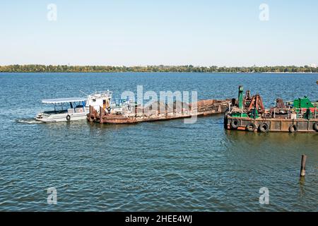 Reinigung der neu entstandenen Insel von Industrieabfällen auf dem Dnjepr mit einem Bagger. Ein Lastkahn mit einem Schlepper nimmt Müll heraus. Ökologisches Konzept. Stockfoto